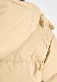Giacca puffer beige con cappuccio, tessuto testurizzato, cuciture a vista e un sottile logo "LACOSTE" di lato.
