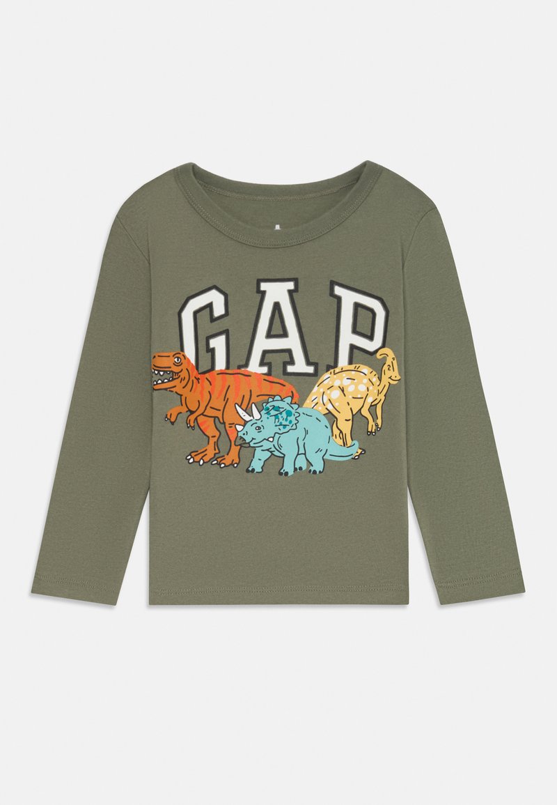 Olīvu zaļš bērnu krekls ar garām piedurknēm, uz kura ir "GAP" teksts un ilustrācijas ar oranžo T-Rex, zilo triceratopsu un dzelteno parasaurolophus.