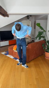 Νέος άνδρας προσαρμόζει φούτερ με κουκούλα ανοιχτό μπλε, στέκεται σε ξύλινο πάτωμα κοντά σε καφέ καναπέ και πράσινο φυτό σε μοντέρνο σαλόνι.