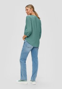 Mintgroene blouse met 3/4 mouwen, gecombineerd met lichtblauwe jeans en mintkleurige sneakers, weergegeven vanaf de achterkant tegen een effen achtergrond.