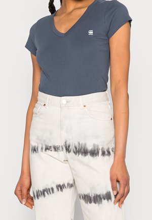 Kvinde iført en tætsiddende mørkeblå V-hals T-shirt med et lille logo og højtaljede, hvide tie-dye jeans med sorte vandrette striber.