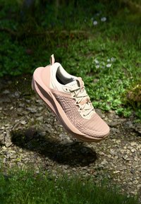 Zapatilla de running ligera en beige con un upper texturizado, una media suela acolchada y un acento rosa en el talón, flotando sobre una superficie de hierba.