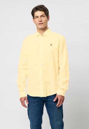 Camicia a maniche lunghe gialla, con bottoni, realizzata in tessuto leggero, con un logo ricamato discreto sul petto, abbinata a jeans blu.