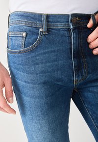 Kék farmernadrág, slim fit szabásban, öt zsebes kialakítással, kontrasztos varrással és közepes mosású felülettel. Elöl gombos és cipzáras záródás.