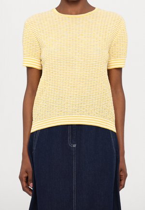 Kobieta ubrana w teksturowany, żółto-biały, prążkowany top z krótkim rękawem, w zestawieniu z ciemnoniebieską spódnicą dżinsową z białym przeszyciem.