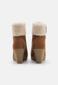 Botas de tornozelo em camurça castanha com um colarinho de pelagem creme, salto em cunha empilhado e fecho lateral para fácil calçamento.