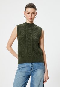 Jersey sin mangas de color verde oliva, tejido, con cuello alto, que presenta un patrón de trenzado texturizado y un dobladillo de canalé, combinado con jeans de color azul claro.