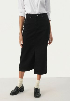 Person iført en sort midikjole, hvid langærmet skjorte med knapper, cremefarvede strømper og sorte loafers, stående mod en ensfarvet baggrund.