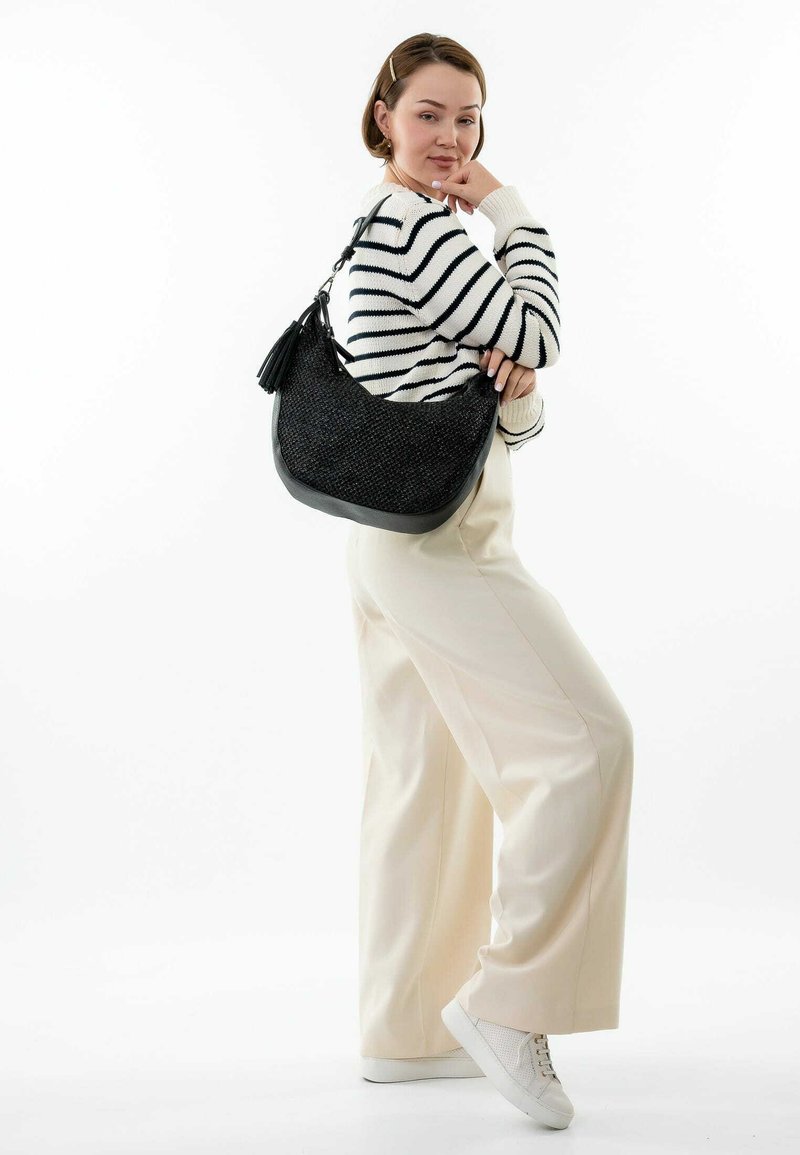 Bolso de mano negro tejido con forma curva y acabado texturizado, que presenta un fondo de cuero y un detalle de borla, llevado sobre un suéter a rayas.