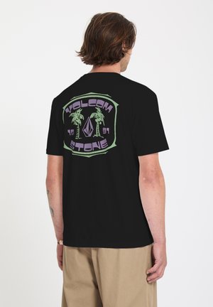 Hombre con camiseta negra de Volcom Stone con logo de palmera verde y morada en la espalda, combinado con pantalones beige, de pie sobre fondo blanco.