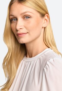 Lyshåret kvinde med langt hår, iført en lysfarvet bluse med en samlet halsudskæring og blød, flydende tekstur, mod en neutral baggrund.