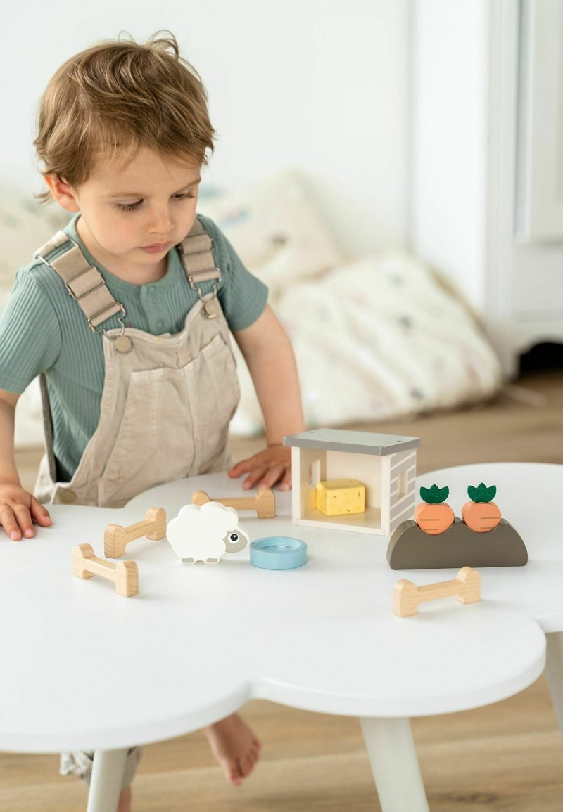 Drewniany plac zabaw zawiera białą owcę, marchewki, niebieską miseczkę oraz dom z serem. Ułożone na białym, okrągłym stole.