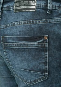 Jeans de mezclilla en azul oscuro con una textura suave, que presentan un parche de logo visible en la cintura y un pequeño bolsillo trasero con un remache de metal.