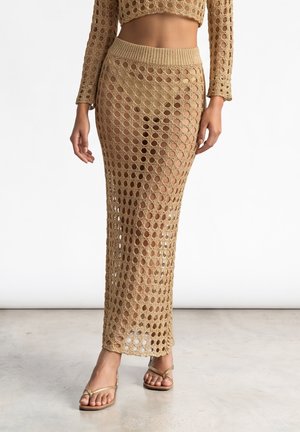 Donna che indossa una gonna maxi beige all'uncinetto e un top corto abbinato a maniche lunghe con sandali con cinturini su uno sfondo semplice.