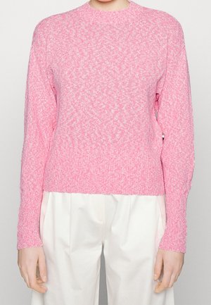 Lichtroze gebreide trui met lange mouwen, gecombineerd met losse witte broek bij een persoon die tegen een effen achtergrond staat.