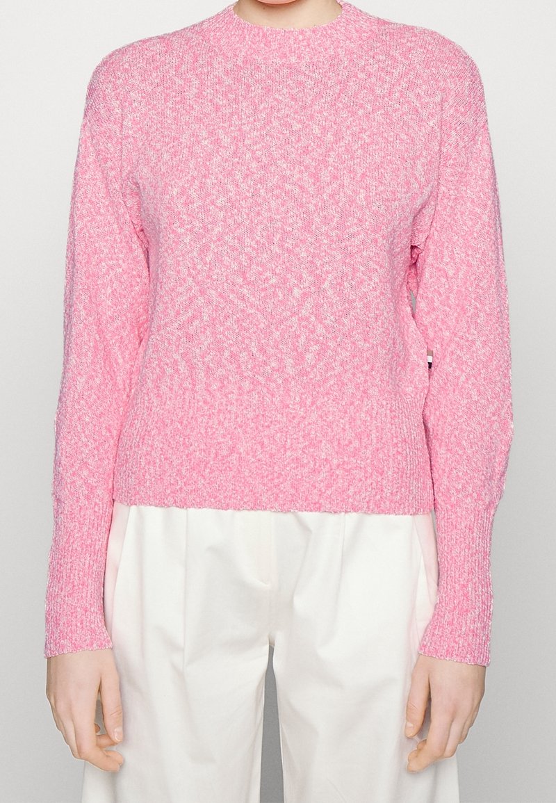 Pull en maille rose clair à manches longues associé à un pantalon blanc ample sur une personne debout devant un fond uni.