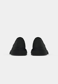Chaussures noires à enfiler avec un dessus en tissu lisse, bout rond, semelle en caoutchouc texturée et design minimaliste sur le panneau arrière.