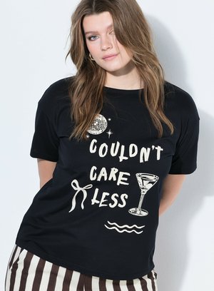 Femme aux longs cheveux châtain clair porte un t-shirt noir avec le texte « Couldn't Care Less », boule disco, verre à martini et motifs de nœud.