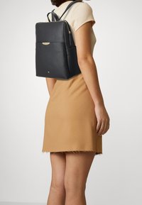 Černý kožený batoh s texturovaným povrchem, zipovým uzávěrem, zlatými kovovými doplňky a vnější kapsou, nošený na rameni.