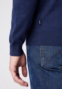 Marinblå stickad tröja med ribbat nederkant och ärmslut, i kombination med blå denimjeans som har ett distinkt sydd mönster på bakfickan.