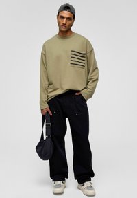 Olijfgroene sweatshirt met zwarte strepen op de borstzak, gecombineerd met losse zwarte broek. Accessoires omvatten een kleine zwarte tas en sneakers.