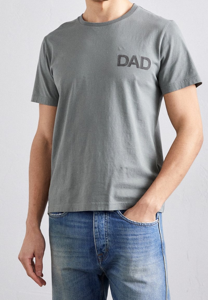 T-shirt en coton gris à manches courtes et col rond, présentant un imprimé "DAD" gris foncé sur la poitrine gauche. Porté avec un jean bleu.