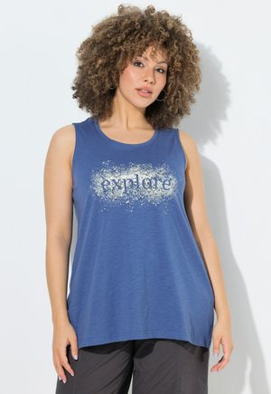 Frau mit lockigem Haar, die ein blaues ärmelloses Oberteil mit dem Schriftzug „explore“ und schwarze Hose trägt, steht vor weißem Hintergrund.