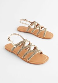 Sandali piatti con cinturini, suola in finta pelle color nocciola e decorazioni in metallo dorato intrecciate. Presentano cinturini alla caviglia regolabili e punte aperte.