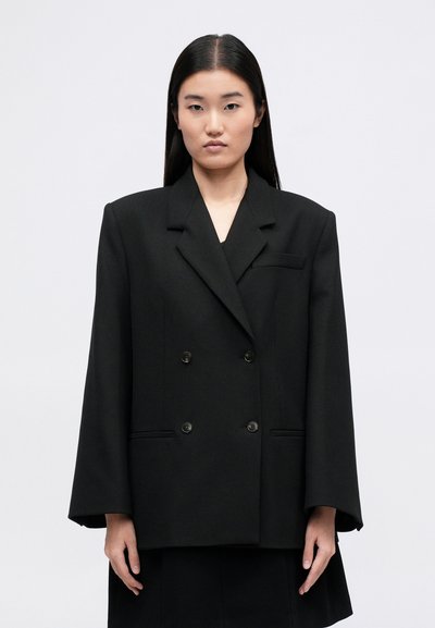 Blazer preto de dupla abotoadura com um design estruturado, apresentando lapelas largas, dois bolsos frontais e tecido texturizado.