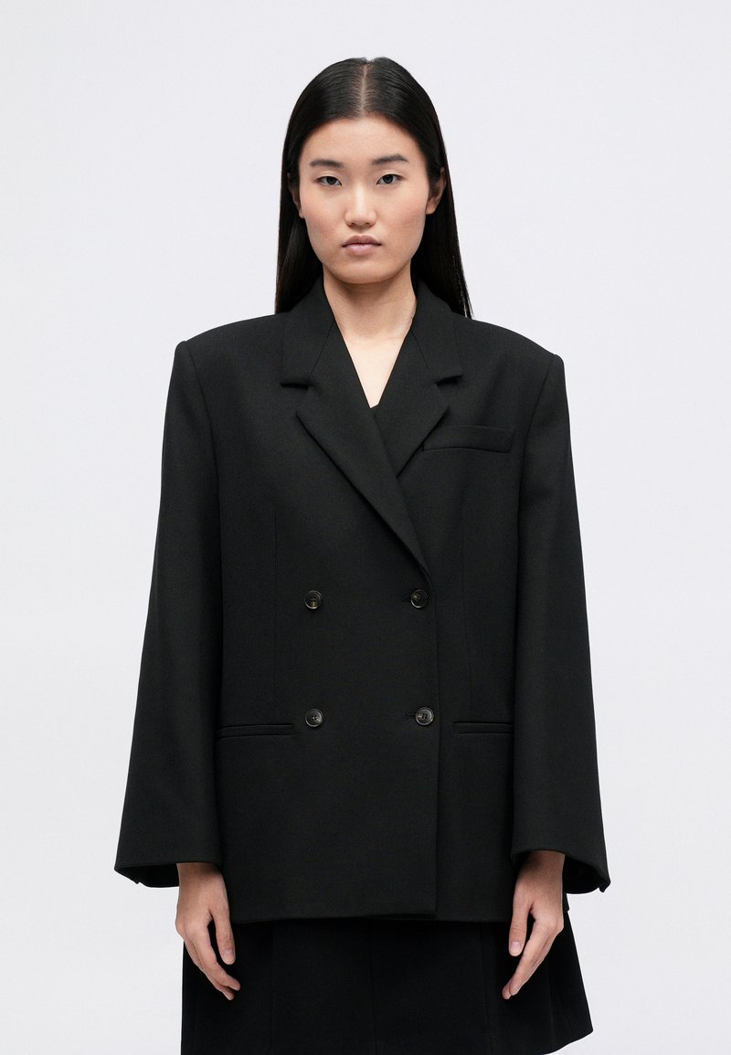 Black double-breasted blazer with a structured design, featuring wide lapels, two front pockets, and textured fabric.
