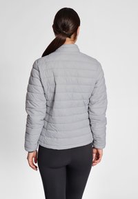 Lys grå polstret jakke med krave, ribbede manchetter og horisontal søm. Bæret over sorte leggings, der viser et simpelt, funktionelt design.