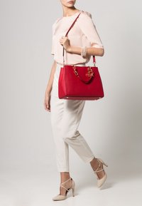 Rote Handtasche mit strukturiertem Design, glatter Textur, goldfarbenen Beschlägen und einer Kettenakzent; getragen zu cremefarbenen Hosen und beigen High Heels.