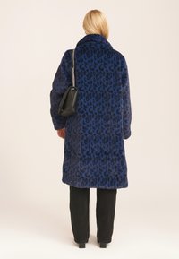 Blue leopard print coat made of soft material, featuring a high collar. Worn over black pants, it is complemented by a black handbag.