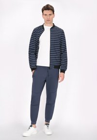 Marine gestreepte bomberjack met geribbelde kraag, witte t-shirt, marine joggingsbroek en witte sneakers met groene accenten.