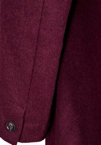 Detail vínové vlněné bundy ukazující jemnou texturu, elegantní střih a černý knoflík. Má boční šev a je dokončena s propracovaným vzhledem.