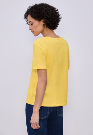 Nainen, jolla on lyhyet kiharat hiukset, pukeutunut yksiväriseen keltaiseen lyhythihaiseen paitaan ja sinisiin farkkuihin, seisoo ja katsoo vasemmalle alas.