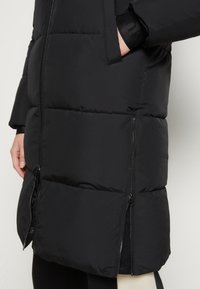 Veste en duvet noir matelassée avec zips à l'avant et sur les côtés, main dans la poche, portée sur un pantalon noir et blanc.