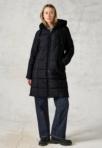 Svart lång pufferjacka med huva, kviltat mönster, dragkedja fram och sidofickor. Bärs över löst sittande mörka jeans och svarta chunky stövlar.