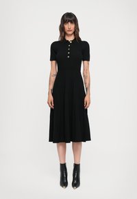 Black ribbed dress with a fitted bodice, short sleeves, and gold buttons; pleated skirt, reaching mid-calf; paired with black pointed ankle boots.