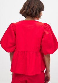 Rode katoenen blouse met een ronde halslijn, pofmouwen en een geraffineerd tailleontwerp. De achterkant toont de vloeiende silhouet en textuur.