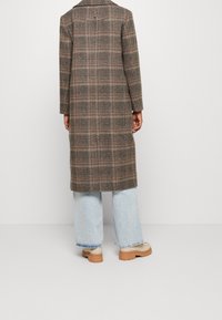 Persoon in een lange bruine geruite jas, lichtblauwe wijde jeans en beige platformschoenen, staande tegen een effen witte achtergrond.