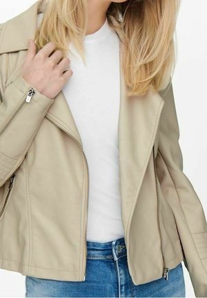 Kvinde iført en beige læderjakke over en hvid skjorte og blå jeans, med den ene hånd let berørende jakkeens krave.