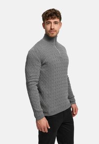 Grå kabelstickad tröja med hög krage och kort dragkedja. Har ribbade muddar och nederkant, med texturerad sömnad över hela.