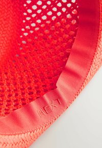 Roter geflochtener Korb mit strukturierter Oberfläche und glattem Satinbesatz. Etikett mit "NEXT", das in den Besatz gestickt ist.