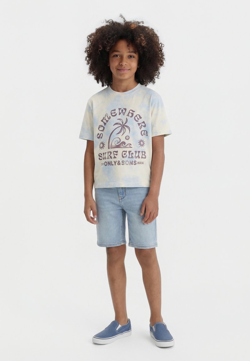 Niño con cabello rizado que lleva una camiseta tie-dye con el texto "Somewhere Surf Club", pantalones cortos de mezclilla clara y zapatos azules tipo slip-on, de pie contra un fondo blanco.