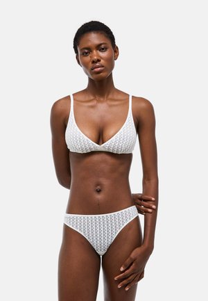 Femme portant un soutien-gorge triangle blanc à motif et un slip taille basse assorti, debout avec un bras dans le dos sur un fond uni.