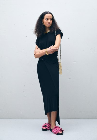 Rochie neagră, ajustată, cu un design răsucit, mâneci scurte. Asortată cu sandale cu detalii florale roz și accesorii din lanțuri aurii, pe un fundal gri.