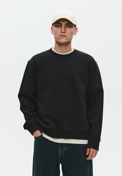 Joven con gorra beige, sudadera negra sobre camiseta blanca y vaqueros oscuros frente a un fondo claro y sencillo.