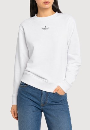 Frau trägt ein weißes Gisada Sweatshirt mit kleinem Logo auf der Brust und blaue Jeans, steht mit einer Hand in der Hosentasche.