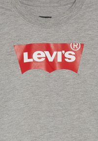 Tricou din bumbac gri, prezentând un mare logo roșu Levi's cu text alb. Logo-ul are forma unui arip de liliac. Textul este centrat pe piept.